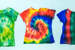 Three tie dye t-shirts on a light background. White clothes painted by hand. Flat lay. Place for text.